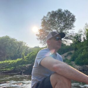 1696680039017 Brandon (author) sitting next to a creek with trees and sun in background.