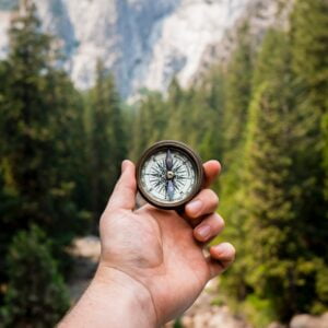 jamie-street-_94HLr_QXo8-unsplash Hand holding a compass in front of a forest of trees, a mountain in the background.