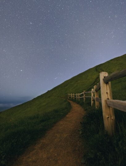 Dirt path leading upwards next to a fence lining a green hill. The path leads to the stars.